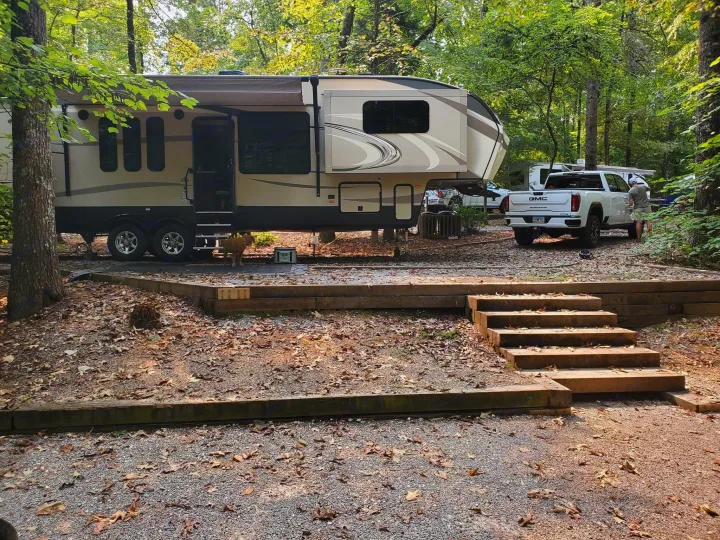 An RV rental setup: a large fifth wheel and a pickup truck parked in a gravel and leaf-strewn wooded campsite. A small dog waits.. Keystone RV Cougar 2017