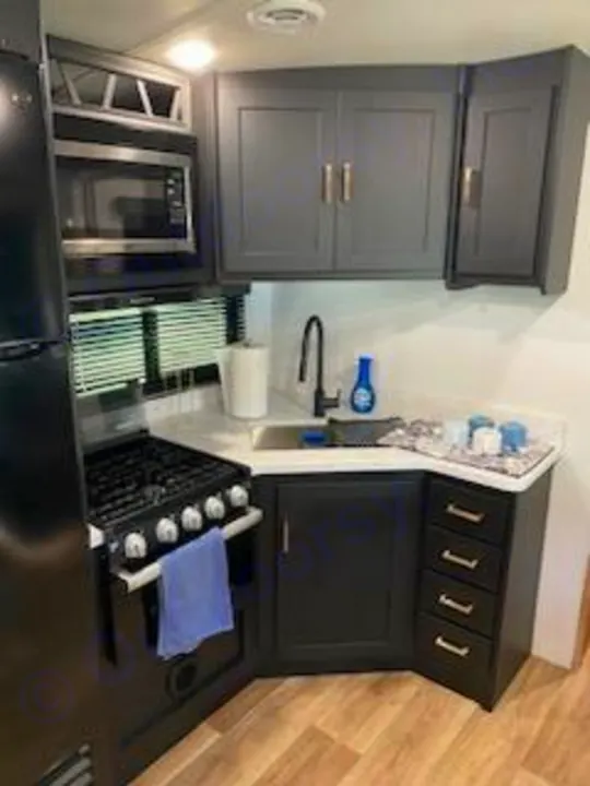 Modern RV rental kitchen: dark gray cabinets, black appliances including a fridge, microwave, and stove, plus a white counter with a sink.. Keystone Outback 2022