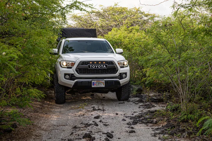 White Toyota Tacoma with rooftop tent, ideal for RV rental adventures, driving on a lush Hawaiian off-road path.. Custom Camper 2016