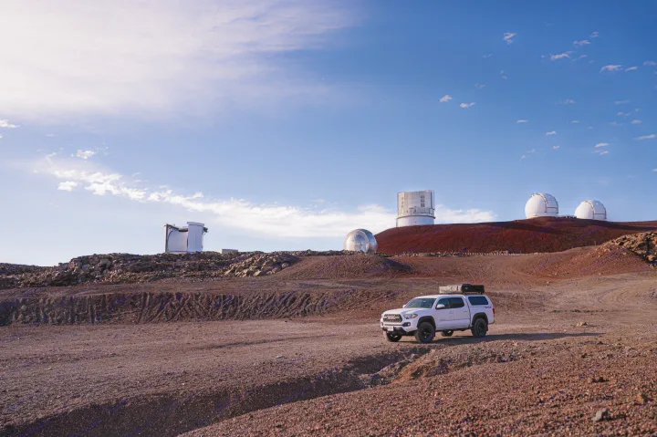 White Toyota Tacoma RV rental exploring Mauna Kea's volcanic landscape near observatories under a bright sky.. Custom Camper 2016