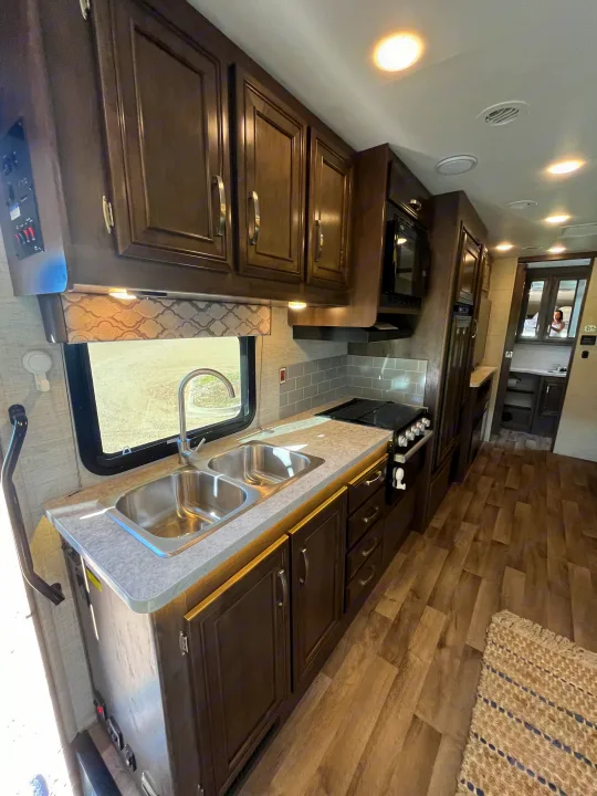 An inviting RV rental kitchen showcasing a double sink, stovetop, microwave, dark wood cabinetry, and wood-look flooring.. Jayco Redhawk 2021