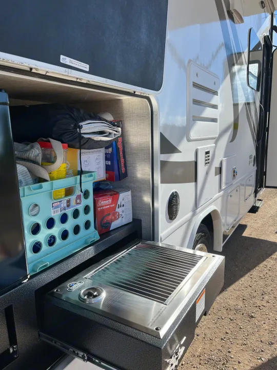 This RV rental shows an open storage bay with camping gear and a convenient pull-out grill for outdoor cooking.. Jayco Redhawk 2021