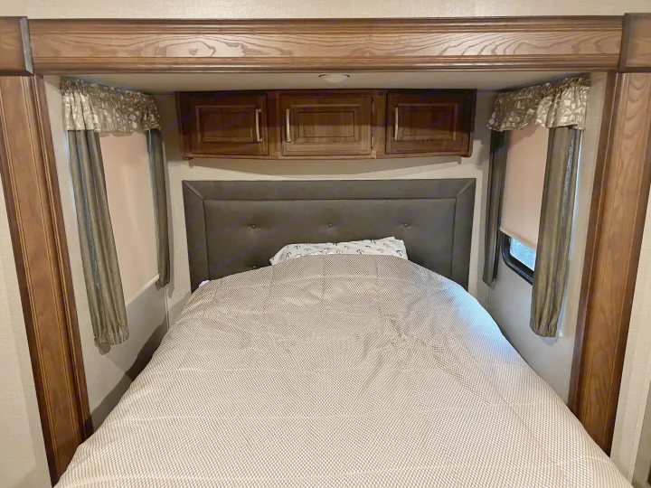 Cozy RV rental bedroom with dark headboard, patterned bedding, overhead cabinets, and window treatments.. Keystone RV Cougar 2017