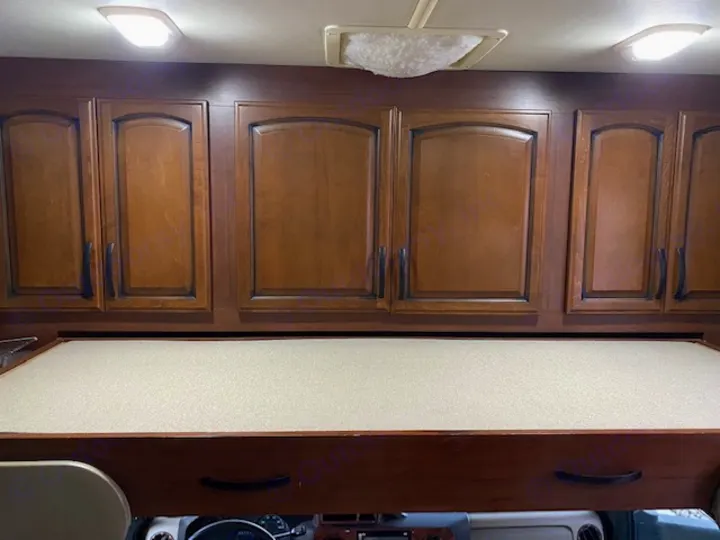 An RV rental interior: wood overhead cabinets with black handles, and a light-colored bunk bed above the cab.. Tioga Fleetwood 2011