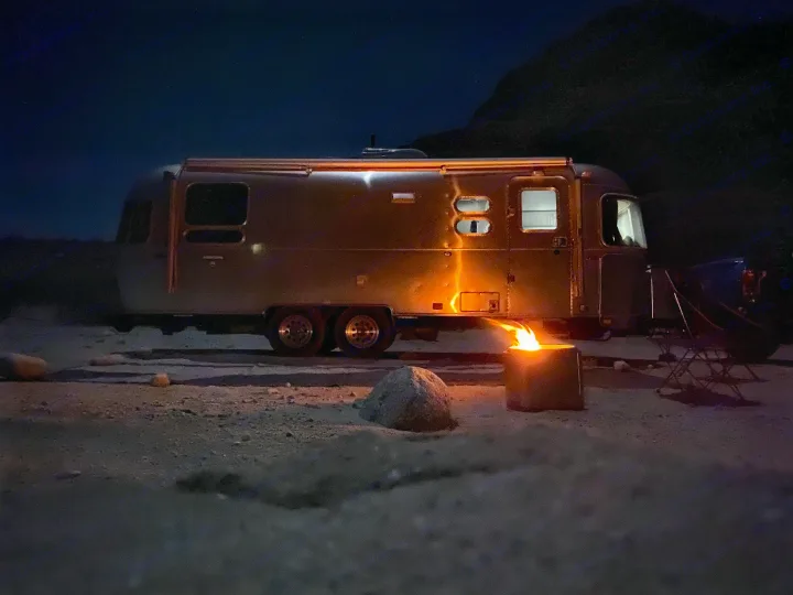 A classic Airstream RV rental glows at night, lit by a crackling campfire, creating a cozy desert camping scene under stars.. Airstream International 2017