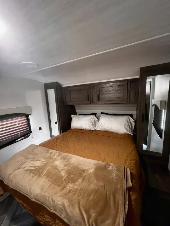 Cozy RV rental bedroom featuring a comfy bed with brown comforter, overhead wood cabinets, and window blinds.. Forest River Evo 2020