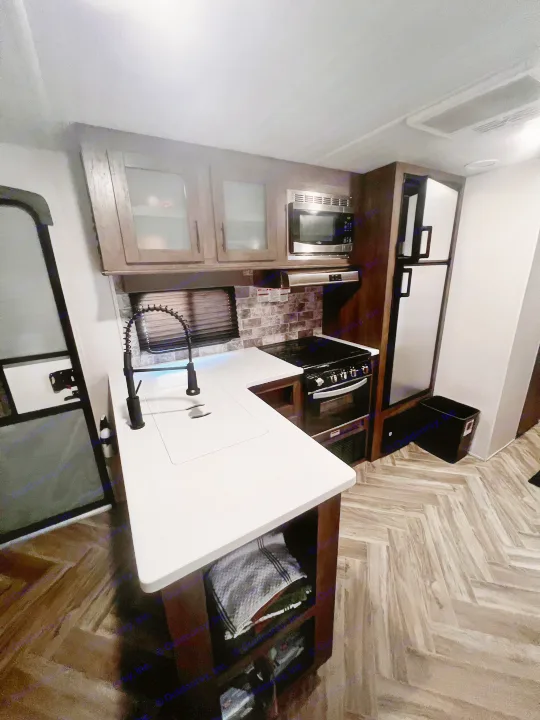 Inside an RV rental: a modern kitchen with white counters, sink, stove, microwave, & fridge on herringbone floors.. Forest River Evo 2020