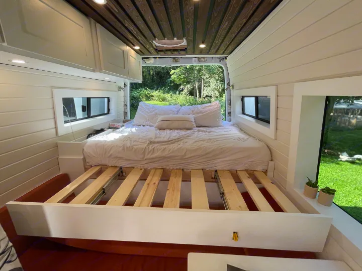 A custom RV rental interior showcasing a cozy bed with pull-out slats, white paneling, and an inviting forest backdrop.. Mercedes Sprinter 2013