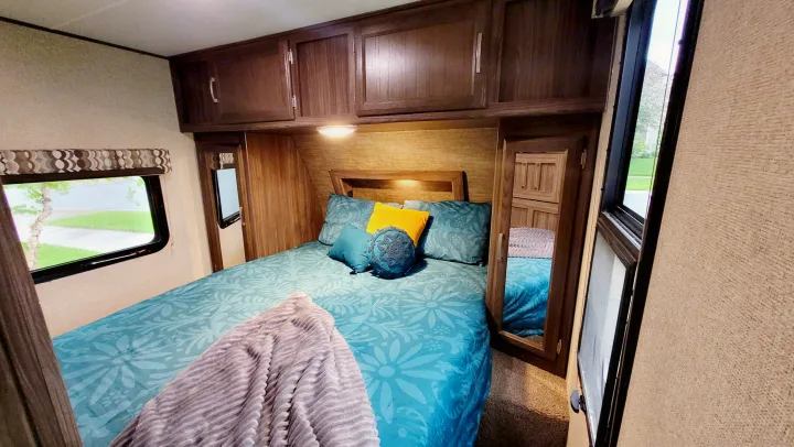 Cozy RV rental bedroom featuring a bed with teal and blue bedding, dark wood overhead cabinets, and an outside window view.. Coachmen Apex 2018