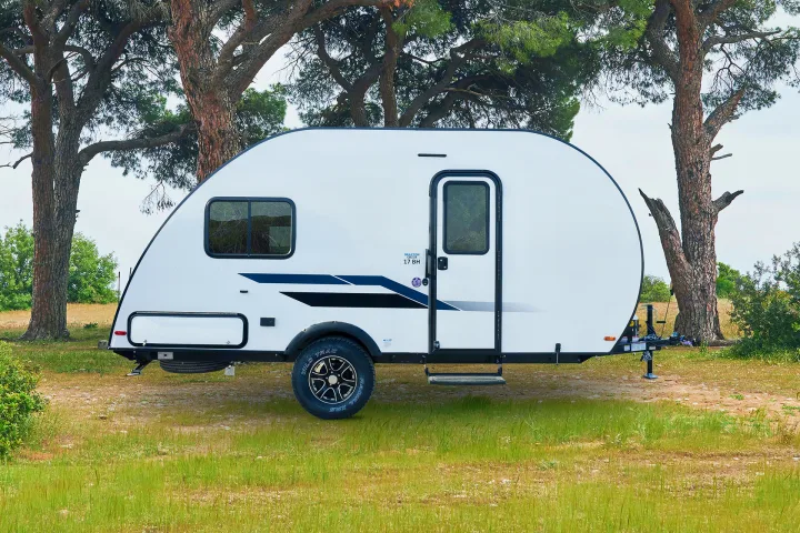 Explore nature with this sleek white rv rental. Compact travel trailer with blue accents, parked among trees in a grassy field.. Braxton Creek BUSHWHACKER PLUS 17FD 2022