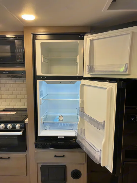 Interior of an RV rental kitchen, featuring an open, empty two-door refrigerator with a thermometer, microwave, and stove.. Jayco Jay Feather 2022