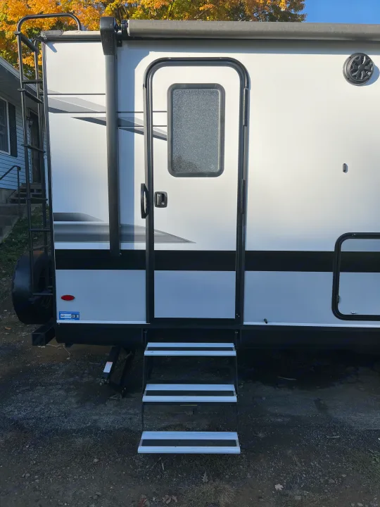 Side of a white RV rental featuring three aluminum entry steps, a main door with a textured window, and a rear roof ladder.. Jayco Jay Feather 2022