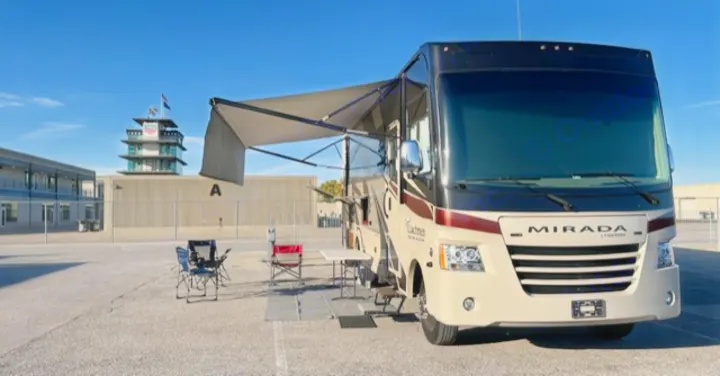 RV rental parked at Indianapolis Motor Speedway with its iconic pagoda tower in the background. Setup has awning, chairs, table.. Coachman Mirada 2018