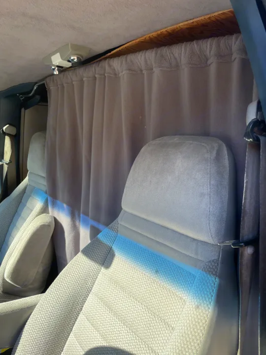 A sunlit view inside an RV rental shows light grey fabric front seats and a pulled privacy curtain, separating the cabin space.. Gmc Custom 1995