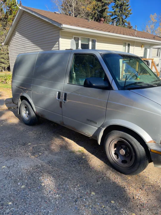 A silver GMC Safari van, a practical choice for a compact RV rental, parked on a sunny gravel driveway by a house.. Gmc Custom 1995