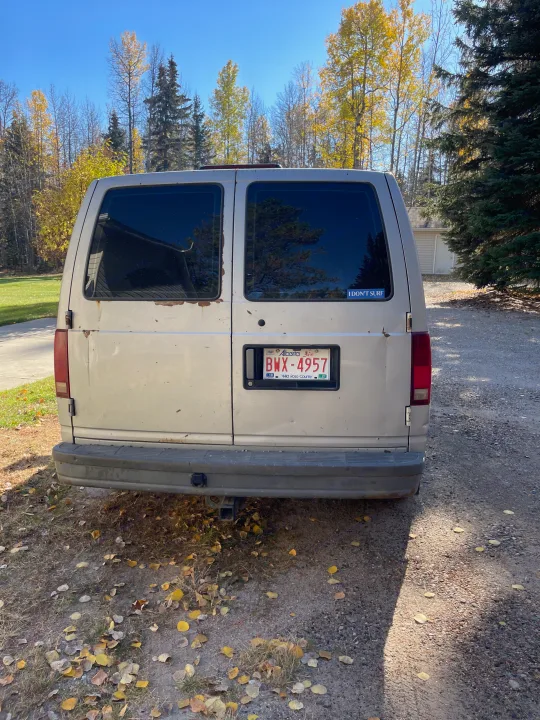 Rear of a beige van, possibly an older RV rental, parked on a gravel drive with autumn leaves.. Gmc Custom 1995