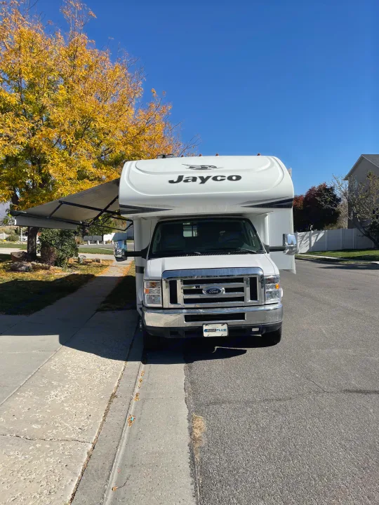 Front view of a white Jayco RV rental with its awning extended, parked on a residential street under a bright autumn sky.. Jayco Greyhawk 2018