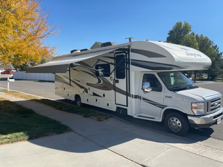 White Class C RV rental parked on a sunny street with awning extended, golden autumn leaves visible.. Jayco Greyhawk 2018