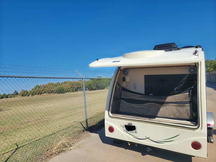 Compact rv rental travel trailer with its rear hatch open, revealing storage. Parked by a fence next to a soccer field.. R-Pod R-Pod Trailer 2015