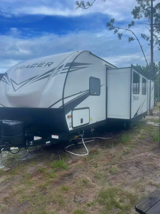 This Tracer RV rental is extended at a peaceful campsite with pine trees under a cloudy sky. Ready for adventure!. Forest River Tracer 2021
