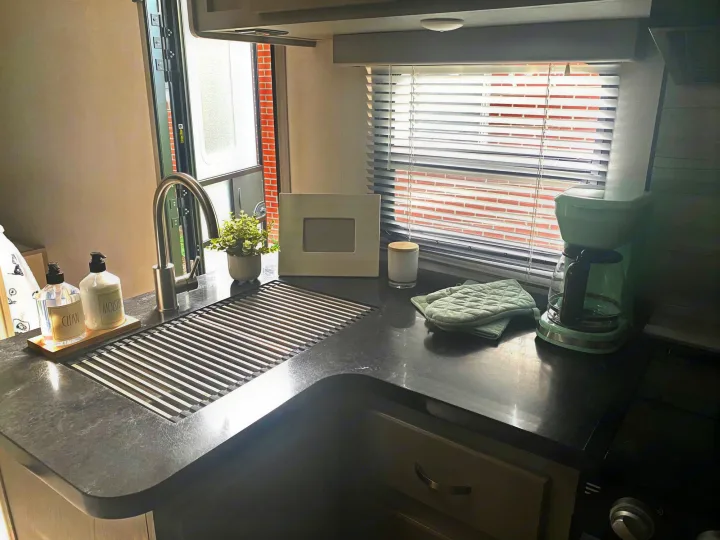 Cozy RV rental kitchen: dark counters, a stainless steel sink with drying rack, light green coffee maker, and a window.. Forest River Tracer 2021