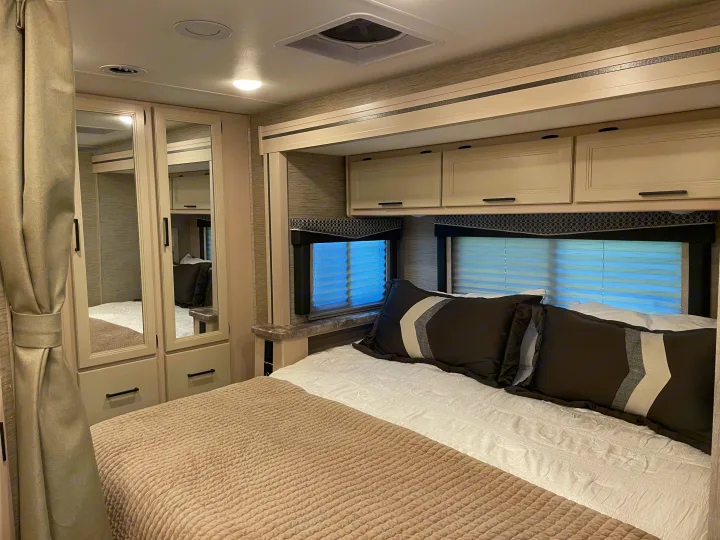 Spacious RV rental bedroom featuring a comfy bed with decorative pillows, mirrored wardrobe, and ample overhead cabinets.. Thor Chateau 2022