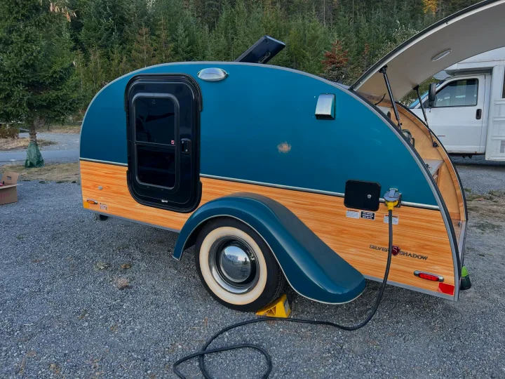 A retro teal and wood teardrop trailer RV rental, a tiny camper, is parked at a campsite with its rear galley open.. Little Guy Silver Shadow 2016
