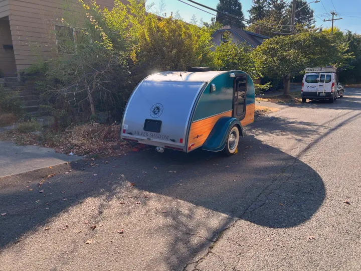 A compact "Silver Shadow" teardrop camper, a type of RV rental, is parked on a sunny residential street.. Little Guy Silver Shadow 2016