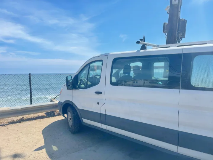 White van, perfect for an RV rental, parked ocean-side. People inside, utility pole nearby. Scenic coastal view.. Ford Transit 2016