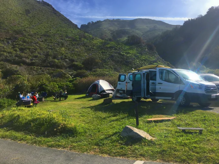 RV rental camper van and tent at a sunny campsite with people, nestled in lush green hills.. Ford Transit 2016