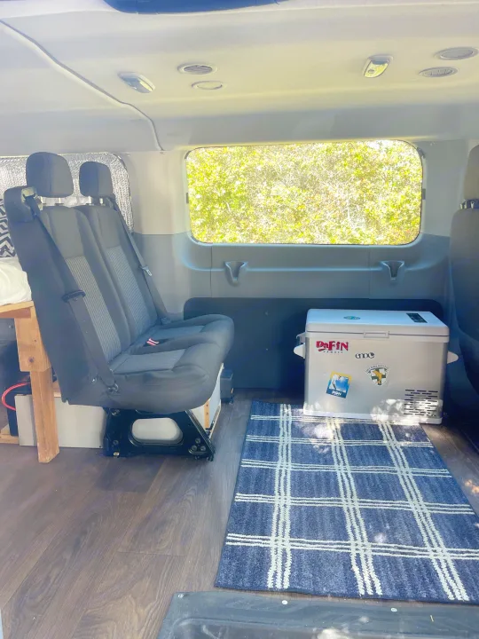 Cozy RV rental interior featuring a two-person bench seat, portable fridge, wood floor, and large window.. Ford Transit 2016