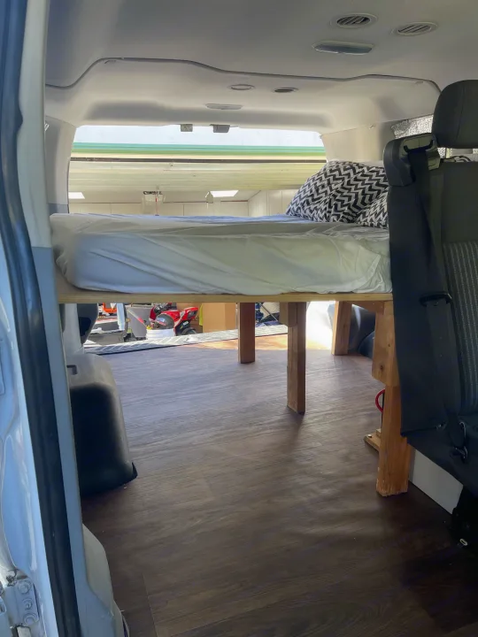 RV rental van's interior: an elevated bed with a white mattress & patterned pillow, offering under-bed storage.. Ford Transit 2016