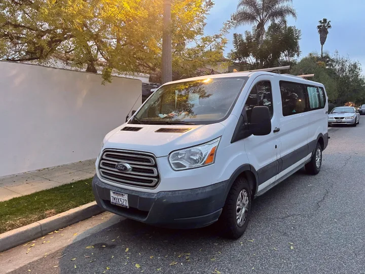 A white Ford Transit passenger van, often used as an rv rental, parked on a tree-lined California street. Plate 7MXK527.. Ford Transit 2016