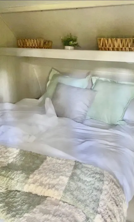 Cozy rv rental bedroom with white sheets, mint green & grey pillows, and a textured blanket. Shelf with baskets and plant adds charm.. Forest River Tracer 2021