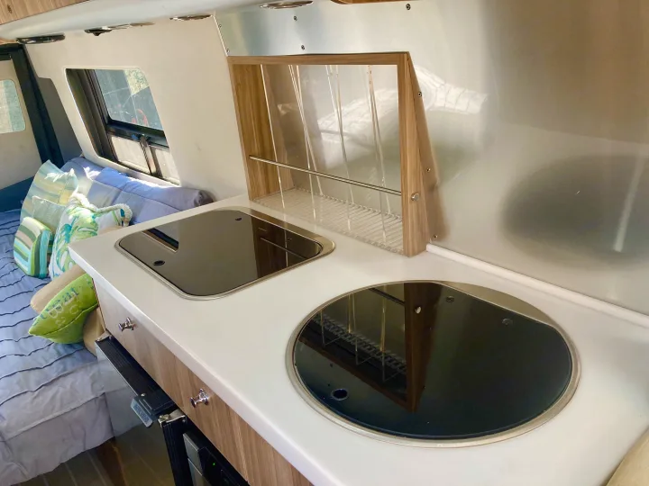 Clean RV rental galley with a compact countertop, covered cooktop and sink, plus an overhead drying rack and bed area.. Airstream Interstate 2017