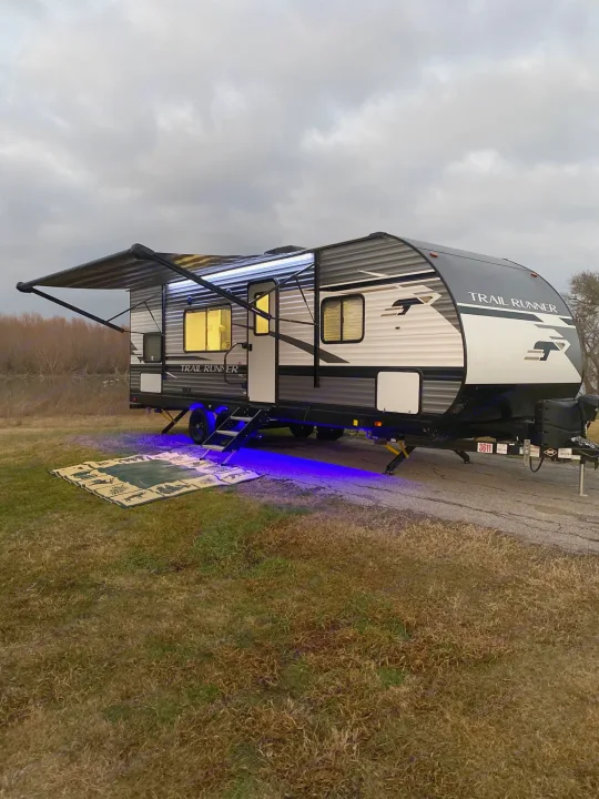 Illuminated RV rental by the water at dusk, featuring an extended awning, glowing interior, blue ground lights, and an outdoor mat.. Heartland Trail Runner 2021