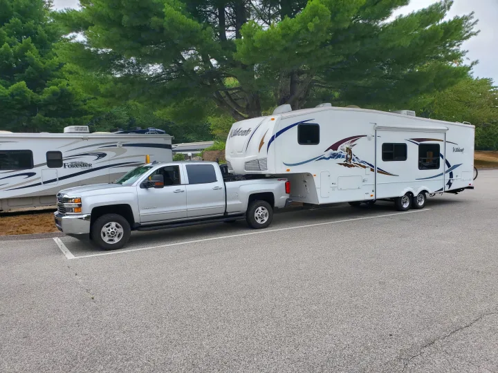 A silver pickup truck tows a large white fifth-wheel RV in a tree-lined lot, ready for an rv rental adventure.. Forest River Wildcat 2009