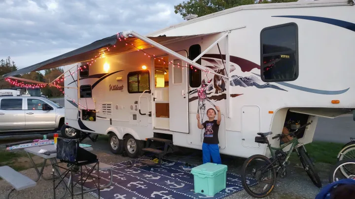 Evening at the RV rental campsite: a fifth-wheel trailer with string lights, a boy with a skeleton, truck, and bikes.. Forest River Wildcat 2009