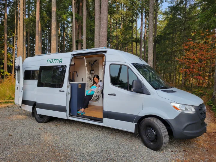 A woman with a baby rests in a hanging chair inside a white NOMA RV rental campervan, parked in a forest.. Mercedes-Benz Sprinter 2021