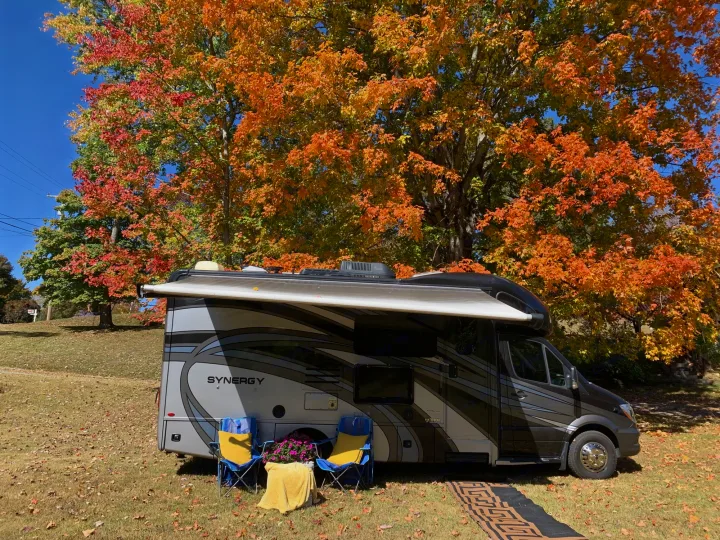 An RV rental, a Synergy model, sits under a tree vibrant with autumn foliage. Camping chairs are set on the grass.. Thor Synergy 2018