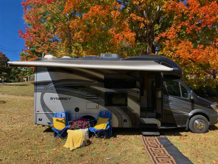 Enjoy an RV rental surrounded by vibrant autumn trees. This motorhome campsite features an awning, chairs, & fall foliage.. Thor Synergy 2018