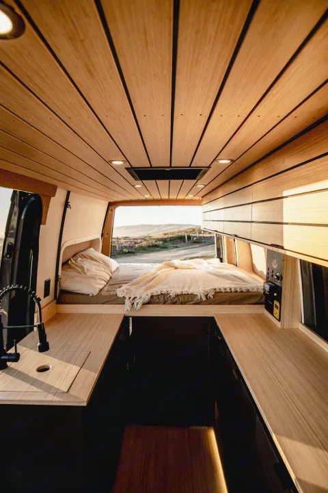 Inside a warm, wooden RV rental: a cozy bed by a scenic window, compact kitchen, and overhead lighting.. Mercedes-Benz Sprinter 2022