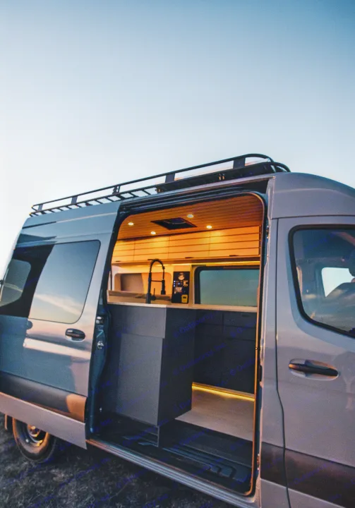 A modern RV rental van under a clear sky, its side door open, shows a warm, lit interior with kitchen amenities.. Mercedes-Benz Sprinter 2022