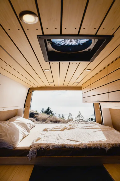 Cozy rv rental camper van interior: a bed, wood ceiling, and panoramic window view of trees and a cloudy sky.. Mercedes-Benz Sprinter 2022