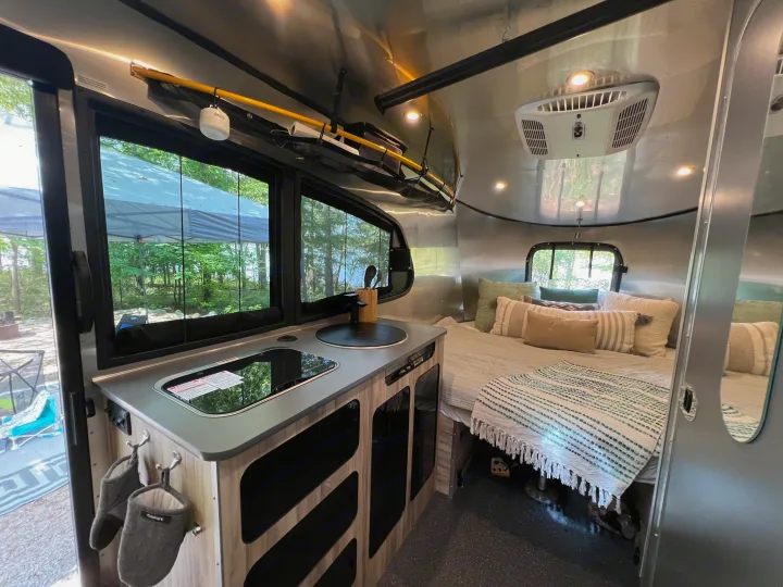 Inside a modern RV rental: sleek kitchen, cozy bed, and large windows revealing a scenic camping spot.. Airstream Basecamp 2022