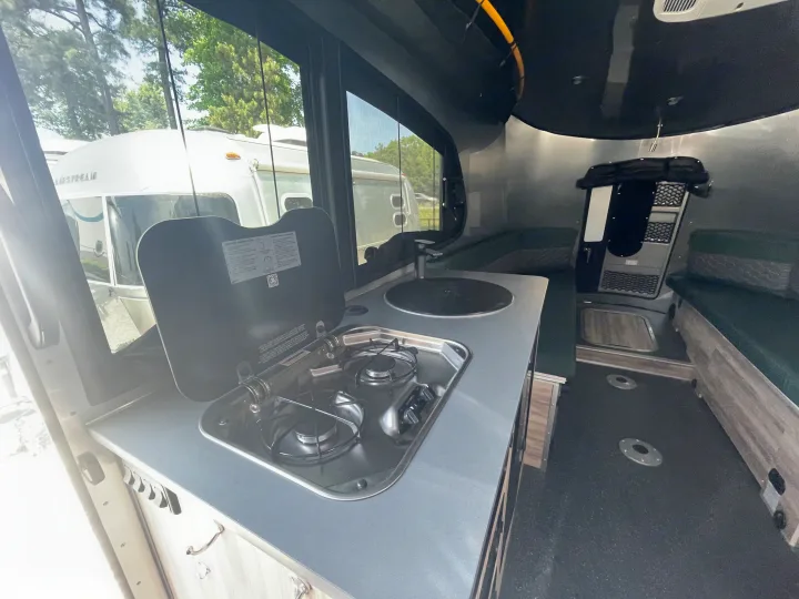 Interior of an RV rental showing a compact kitchenette with stove, sink, and green seating. Another Airstream camper is visible outside.. Airstream Basecamp 2022