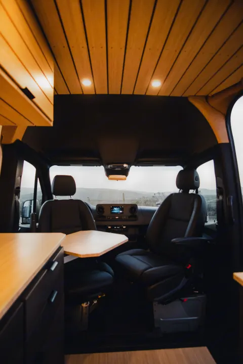 This RV rental features a modern van interior with swivel seats, a table, and warm wooden ceiling lights, perfect for travel.. Mercedes-Benz Sprinter 2022