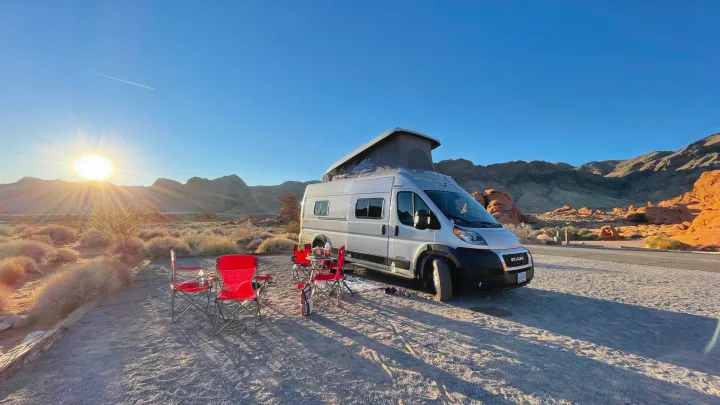 RV rental camper van set for desert camping at sunset. Red chairs, pop-top van, and scenic red rock mountains create an inviting outdoor scene.. Winnebago Solis 2022