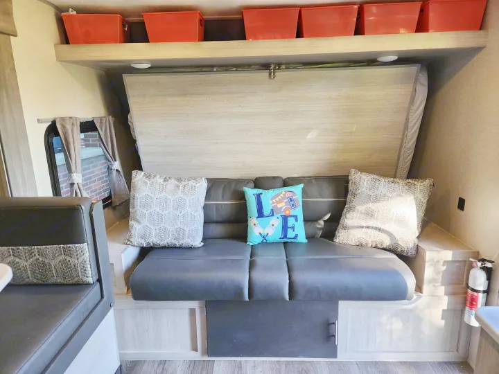 Cozy RV rental interior with a dark couch, decorative pillows, fold-up bed, and overhead storage bins. Fire extinguisher mounted.. Forest River Coachmen Catalina Summit 2022