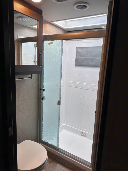 RV rental bathroom with white toilet, a shower with textured glass sliding door, and overhead skylight, designed for travel comfort.. Grand Design Reflection 2022